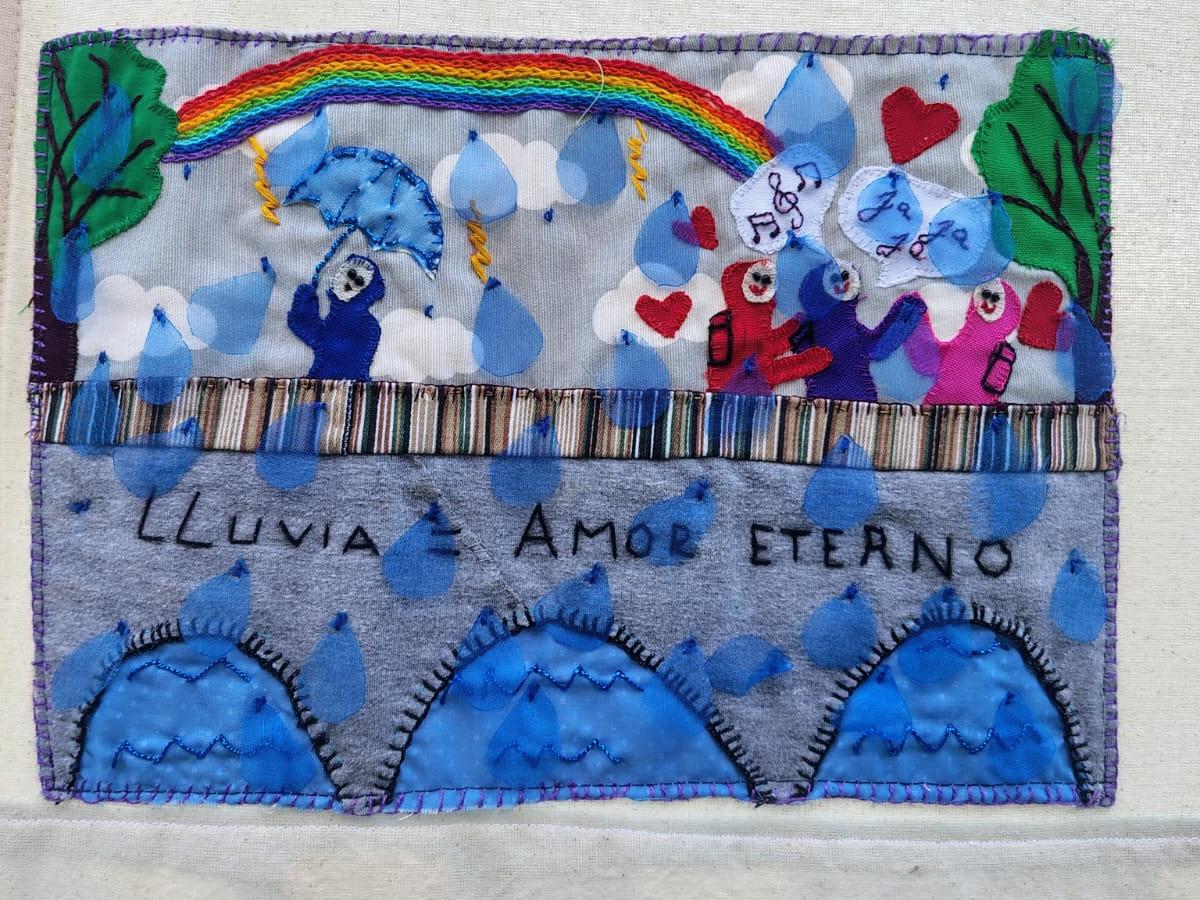 Arpillera colorida que representa una escena bajo la lluvia, con personas sosteniendo paraguas, un arcoíris en el cielo y gotas de agua sobre un paisaje; en el centro se lee la frase “Lluvia = amor eterno”.