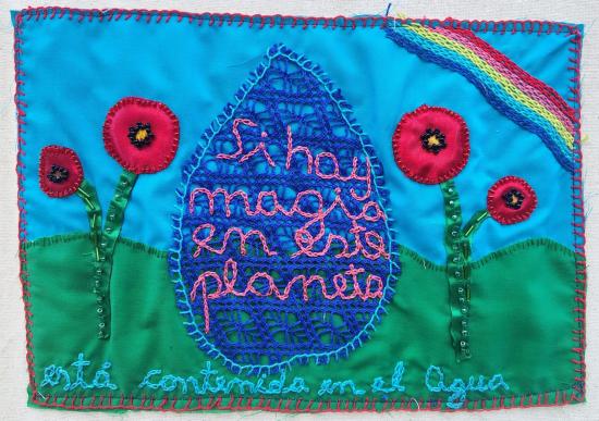 Pancarta textil sostenida en una estructura de madera, compuesta por múltiples bordados de colores que representan paisajes y escenas vinculadas al agua. En el centro se lee el mensaje “Nuestros cauces, nuestra causa”.